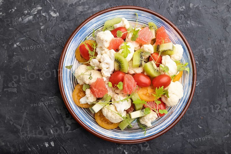 Vegetarian salad of cauliflower cabbage, kiwi, tomatoes, microgreen sprouts on black concrete background. Top view, close up