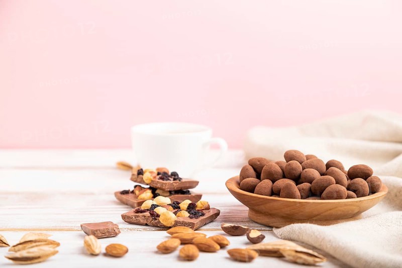 Almond in chocolate dragees in wooden plate and a cup of coffee on white and pink background and linen textile. Side view, copy space, selective focus