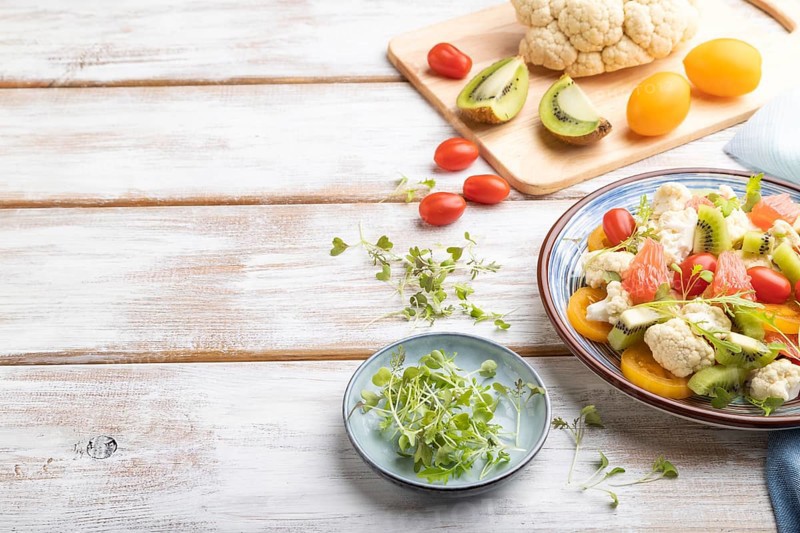 Vegetarian salad of cauliflower cabbage, kiwi, tomatoes, microgreen sprouts on white wooden background and blue linen textile. Side view, copy space