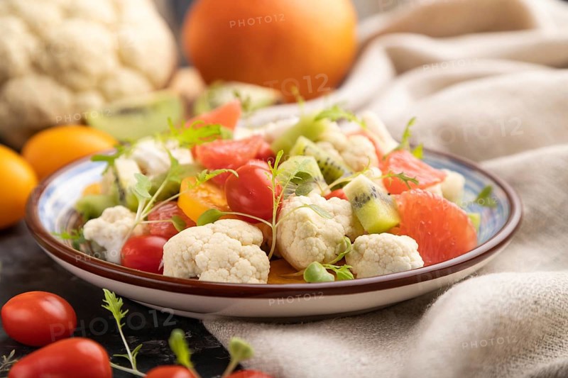 Vegetarian salad of cauliflower cabbage, kiwi, tomatoes, microgreen sprouts on black concrete background and linen textile. Side view, close up, selective focus