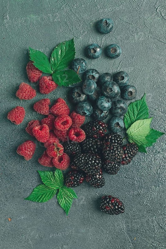 A mix of ripe berries, with leaves scattered on the table, raspberries, blackberries, blueberries, food background