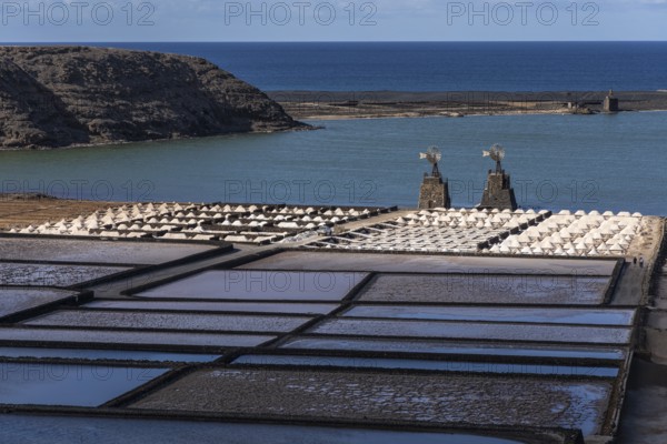 View of salt fields on the coast with blue sea and a barren landscape in the background, two windmills, Canary Islands, Lanzarote, Spain, Europe
