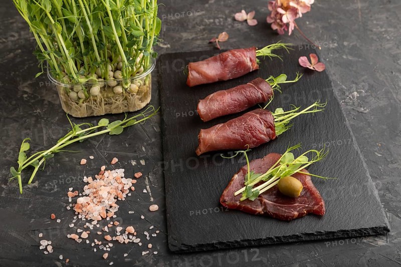 Slices of smoked salted meat with green pea microgreen on black concrete background. Side view, close up