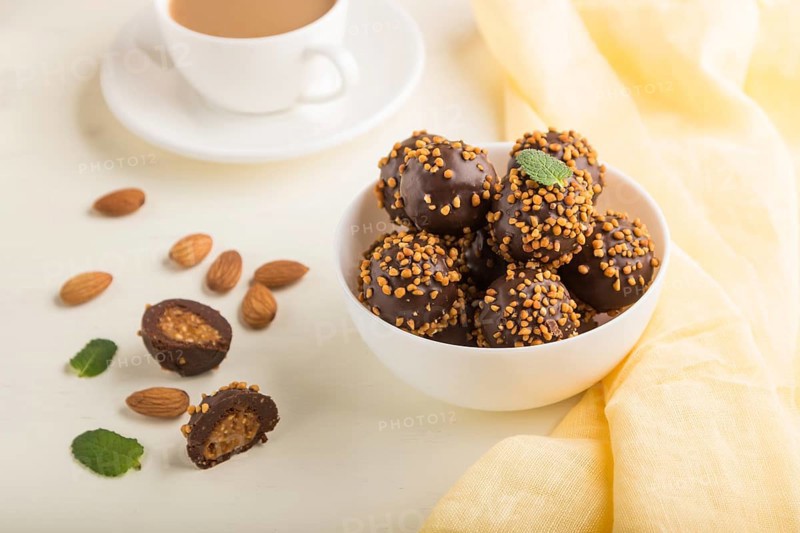 Chocolate caramel ball candies with almonds and a cup of coffee on a white wooden background and yellow textile. Side view, close up, selective focus