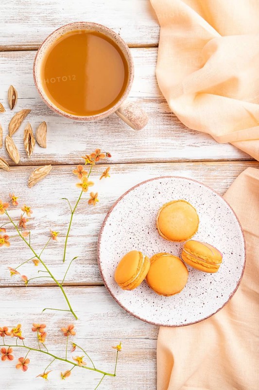 Orange macarons or macaroons cakes with cup of apricot juice on a white wooden background and orange linen textile. Top view, flat lay, close up