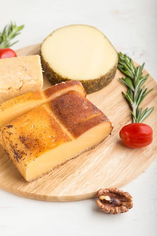 Smoked cheese and various types of cheese with rosemary and tomatoes on wooden board on a white wooden background. Side view, close up, selective focus