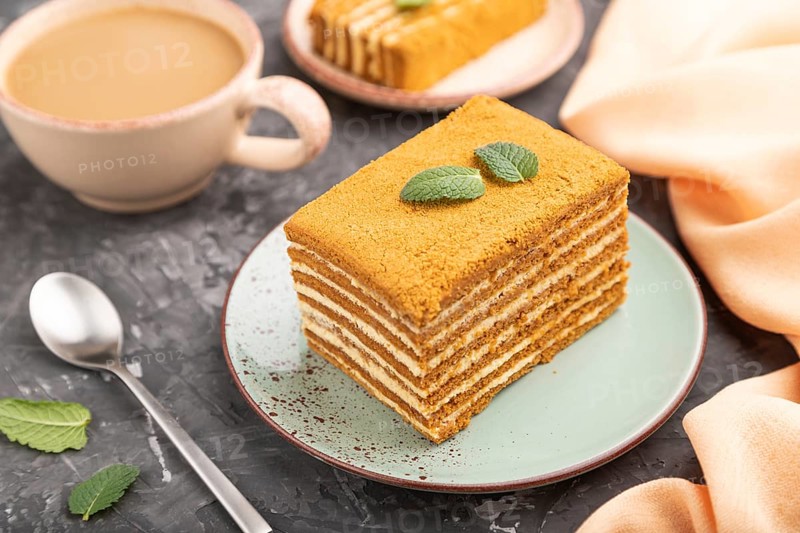 Homemade honey cake with milk cream and mint with cup of coffee on a black concrete background and orange textile. Side view, close up, selective focus