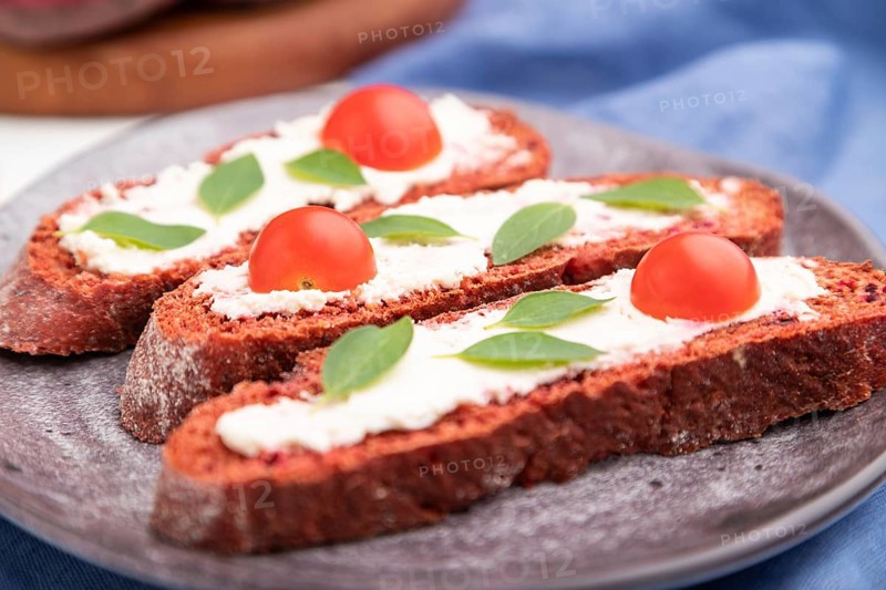 Red beet bread sandwiches with cream cheese and tomatoes on white concrete background and blue linen textile. Side view, selective focus, close up