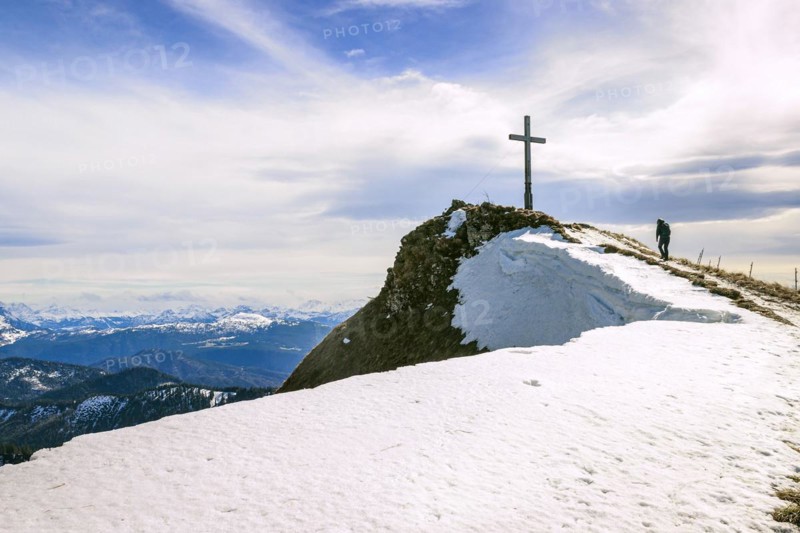 A hiker walks to the Hochgern summit cross over a snow-covered ridge, in the Chiemgau Alps in Bavaria Germany Europe