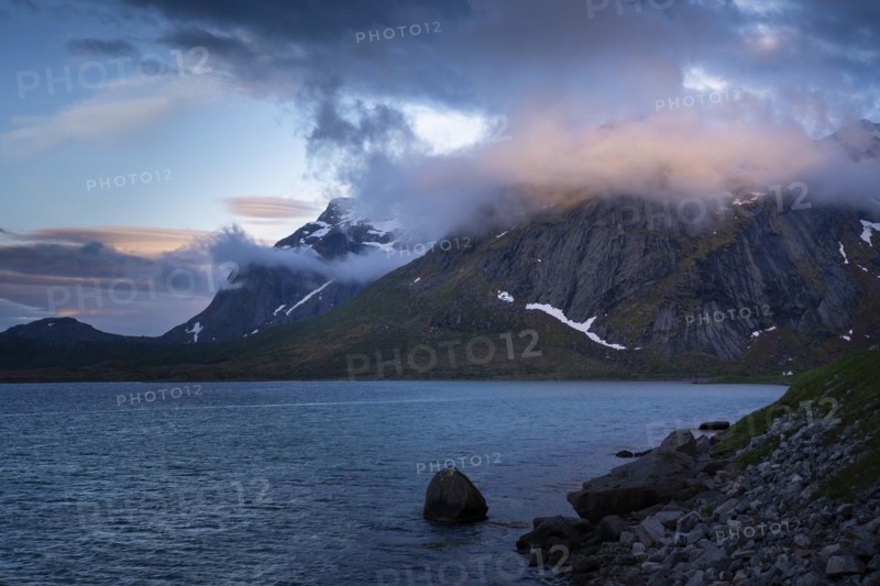 Mountain landscape on the Lofoten Islands. View over the sea to a mountain, the summit is in the clouds, the clouds glow in the sun. At night at the time of the midnight sun. Flakstadoya, Lofoten, Norway, Europe
