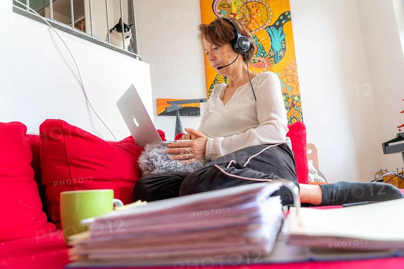 Woman, mid-50s, works from home, with laptop and communicates with colleagues via headset, home office, on the sofa