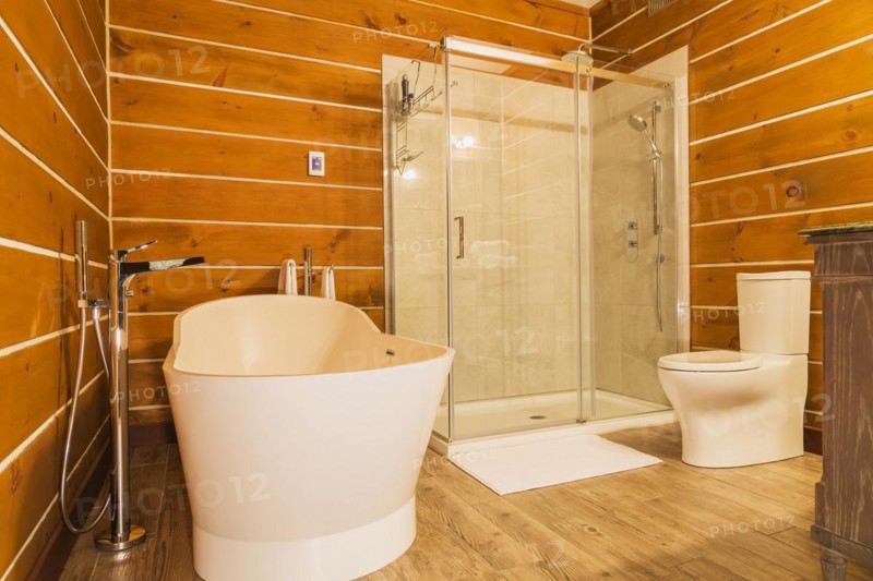 Freestanding bathtub with high tank toilet and clear glass shower stall in en suite with imitation wood ceramic tile flooring inside a milled Eastern white pine timber and flat log profile home, Quebec, Canada, North America