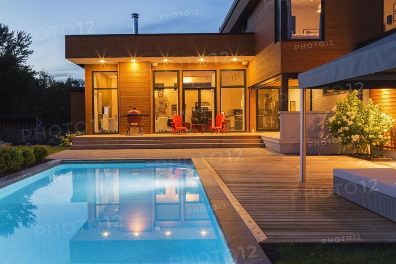 Rear of modern cube style home with stained horizontal wood cladding, in-ground swimming pool and wooden deck with gazebo canopy at dusk, Quebec, Canada, North America