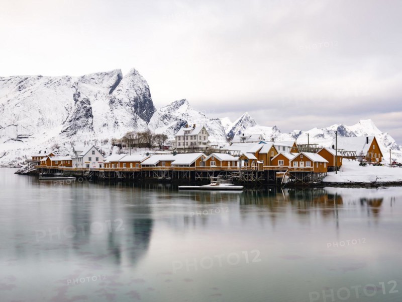 Traditional yellow rorbuer huts on the island of Sakrisøy, snow-covered rocky mountains in the background, Sakrisoy, Reine, Moskenesøya, Lofoten, Norway, Europe