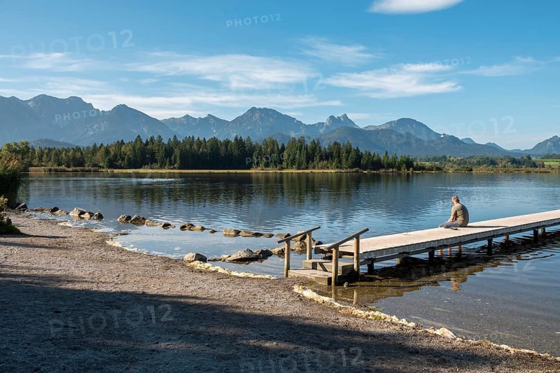 Person sitting on a wooden footbridge looking at a calm lake with mountains in the background, Hopfensee, Hopfen am See, near Füssen, Allgäu Alps, Ostallgäu, Allgäu, Bavaria, Germany, Europe