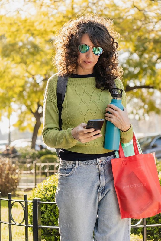 Woman with curly hair and sunglasses uses her smartphone in a park at sunset. Dressed casually in a green sweater and jeans, she holds a water bottle and a red bag, enjoying the outdoor environment