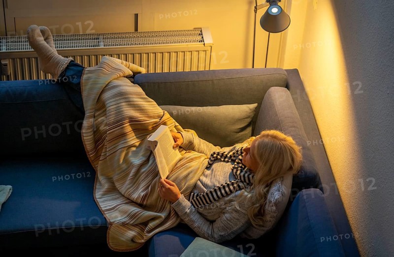 Symbolic image energy saving, cold winter, energy crisis, cold flat, mother and daughter lying on the sofa, warmly dressed, candlelight, heating on to a minimum, warming their feet on the radiator