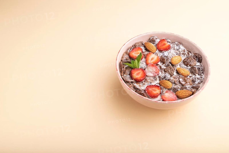Chocolate cornflakes with milk, strawberry and almonds in ceramic bowl on pastel orange background. Side view, copy space