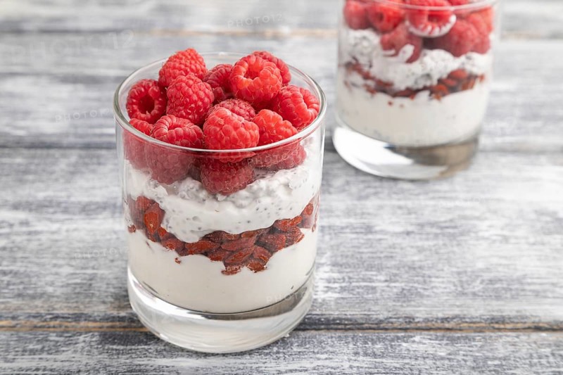 Yogurt with raspberry, goji berries and chia seeds in glass on gray wooden background. Side view, close up, selective focus