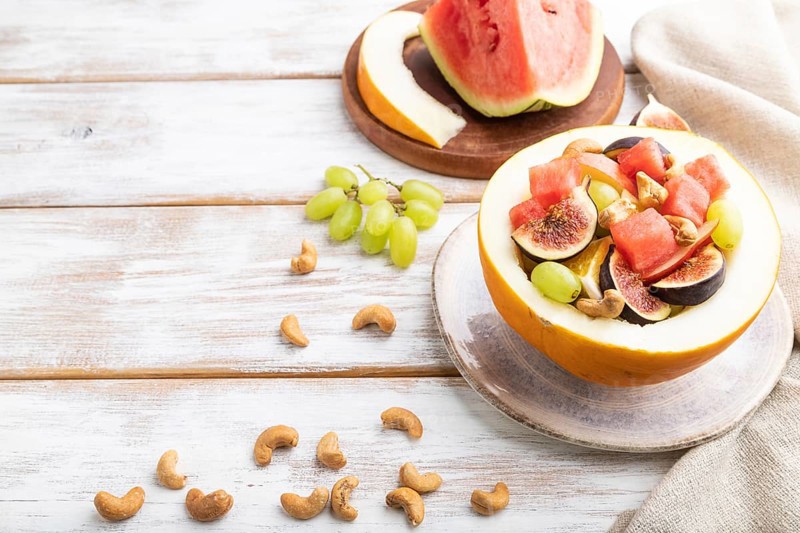Vegetarian fruit salad of watermelon, grapes, figs, pear, orange, cashew on white wooden background and linen textile. Side view, copy space