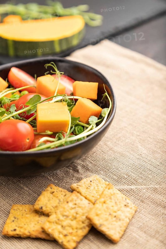 Vegetarian vegetable salad of tomatoes, pumpkin, microgreen pea sprouts on black concrete background and linen textile. Side view, close up, selective focus