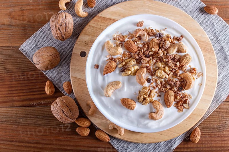 White plate with greek yogurt, granola, almond, cashew, walnuts on brown wooden background. top view