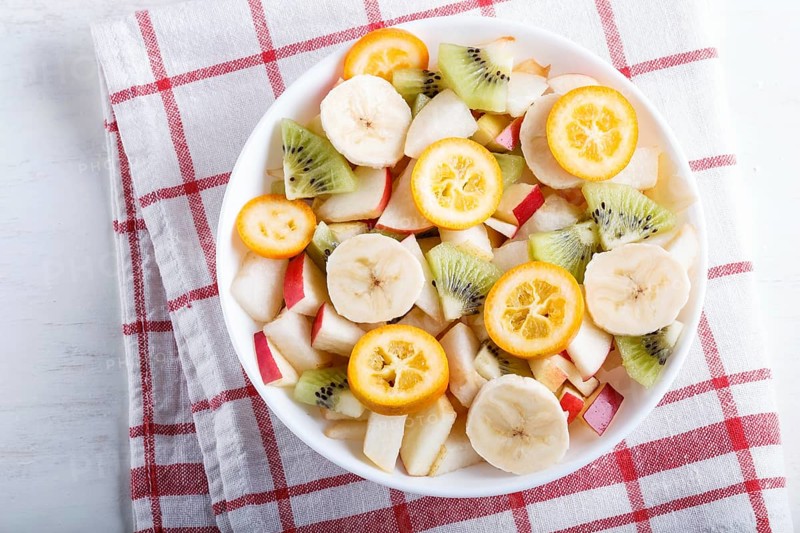 Vegetarian salad of bananas, apples, pears, kumquats and kiwi on linen tablecloth, top view, flat lay, white wooden background