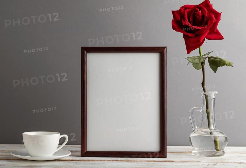 Brown wooden frame mockup with red rose in glass and white coffee cup on gray paper background. Blank, vertical orientation, still life, copy space