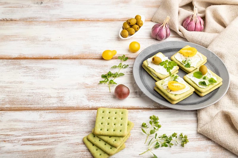 Green cracker sandwiches with cream cheese and cherry tomatoes on white wooden background and linen textile. side view, copy space
