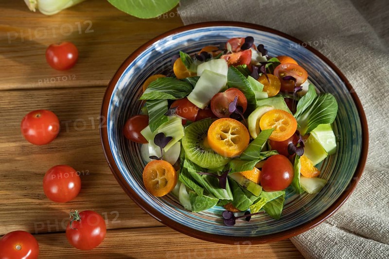 Vegetarian salad of pac choi cabbage, kiwi, tomatoes, kumquat, microgreen sprouts on a wooden background and linen textile. Hard light, contrast. Side view, close up