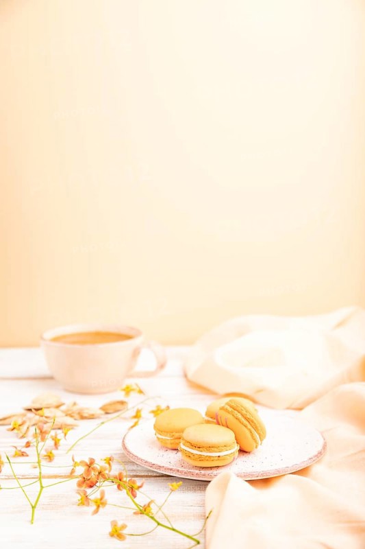 Orange macarons or macaroons cakes with cup of apricot juice on a white wooden background and orange linen textile. Side view, copy space, selective focus