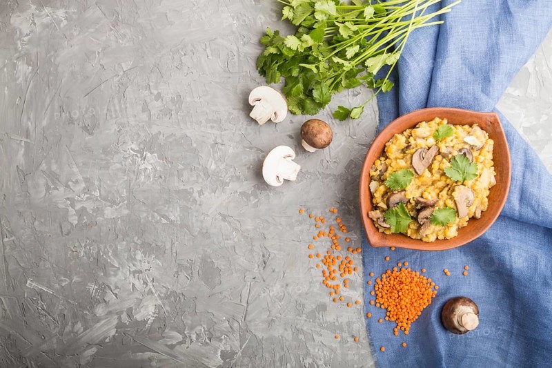 Lentils porridge with champignons and coriander in a clay bowl on a gray concrete background and blue linen textile. Top view, flat lay, copy space