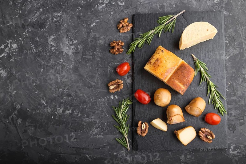 Smoked cheese and various types of cheese with rosemary and tomatoes on black slate board on a black concrete background. Top view, copy space, flat lay