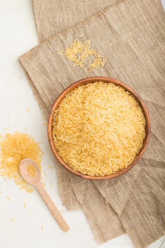 Wooden bowl with raw golden rice and wooden spoon on a white wooden background and linen textile. Top view, flat lay, close up