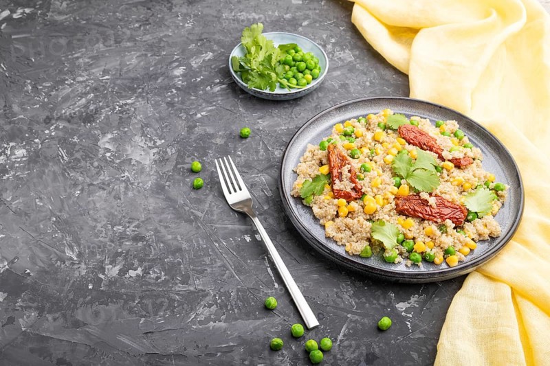 Quinoa porridge with green pea, corn and dried tomatoes on ceramic plate on a gray concrete background and yellow textile. Side view, copy space