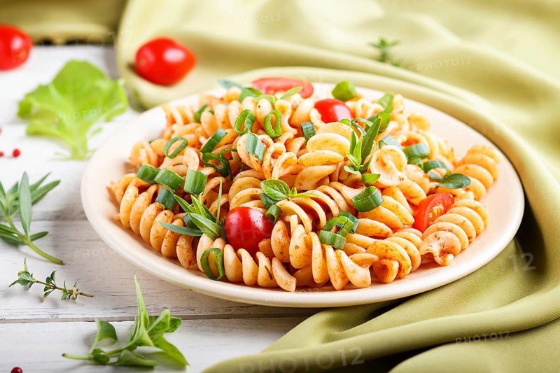 Fusilli pasta with tomato sauce, cherry tomatoes, lettuce and herbs on a white wooden background with green textile. side view, close up, selective focus