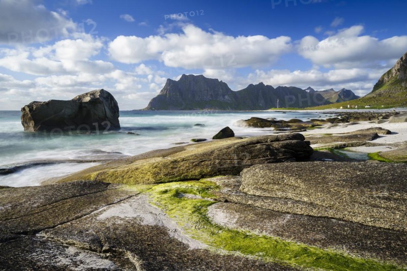 Seascape on the beach at Uttakleiv (Utakleiv), rocks and green seaweed in the foreground. In the background the mountain Hogskolmen. Good weather, blue sky with some clouds. Early summer. Long exposure. Uttakleiv, Vestvagoya, Lofoten, Norway, Europe