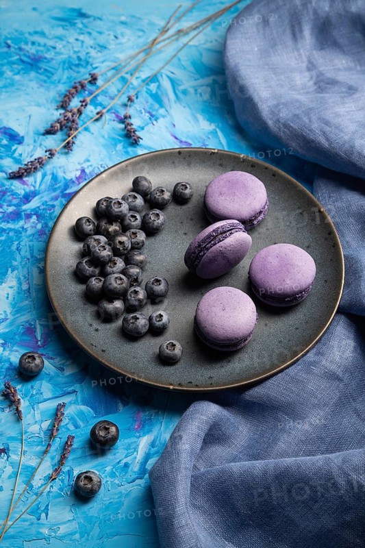 Purple macarons or macaroons cakes with blueberries on ceramic plate on a blue concrete background and blue textile. Side view, close up