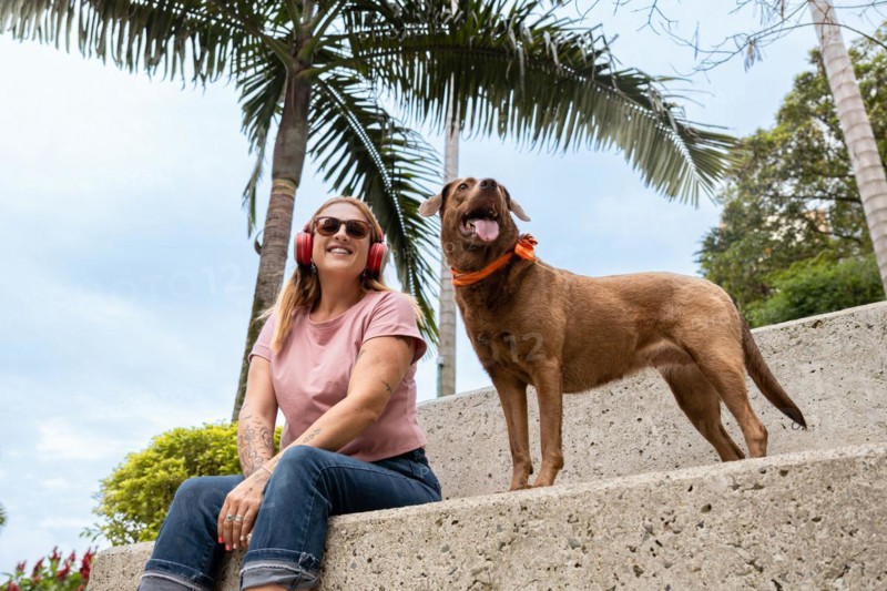 A smiling woman listens to music while sitting next to her alert dog wearing a colorful bandana