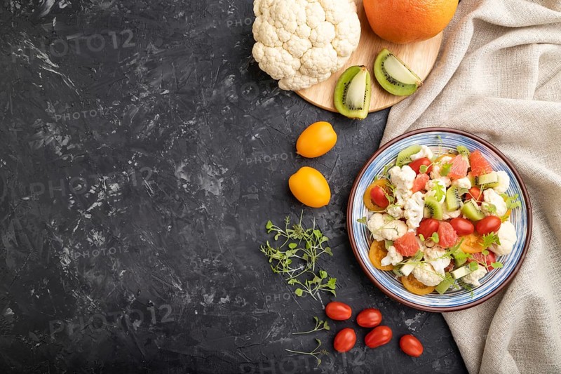 Vegetarian salad of cauliflower cabbage, kiwi, tomatoes, microgreen sprouts on black concrete background and linen textile. Top view, flat lay, copy space