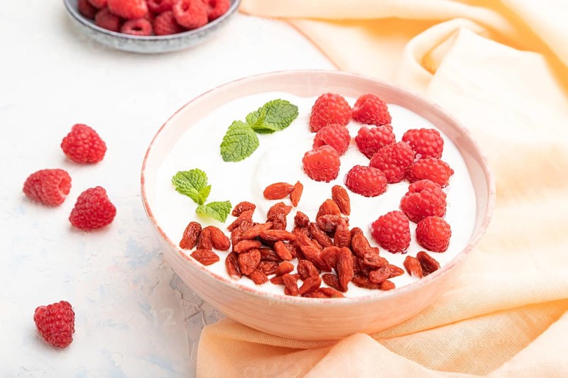Yogurt with raspberry and goji berries in ceramic bowl on white concrete background and orange linen textile. Side view, close up