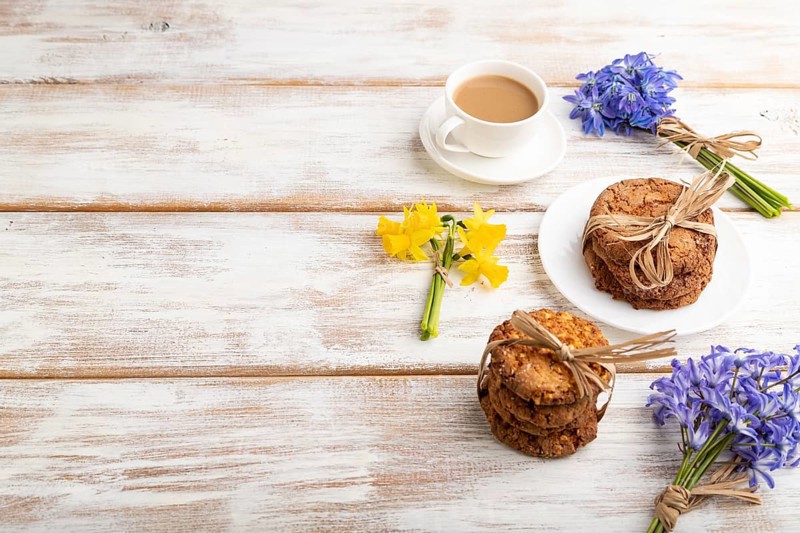 Oatmeal cookies with spring snowdrop flowers bluebells, narcissus and cup of coffee on white wooden background. side view, copy space, still life. Breakfast, morning, spring concept