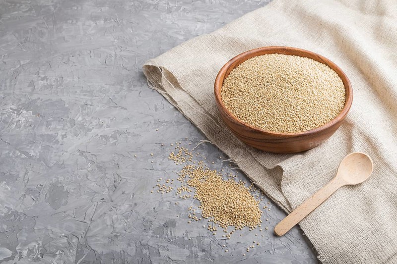Wooden bowl with raw white quinoa seeds and wooden spoon on a gray concrete background and linen textile. Side view, copy space