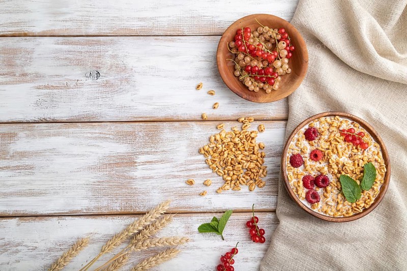 Wheat flakes porridge with milk, raspberry and currant in wooden bowl on white wooden background and linen textile. Top view, flat lay, copy space