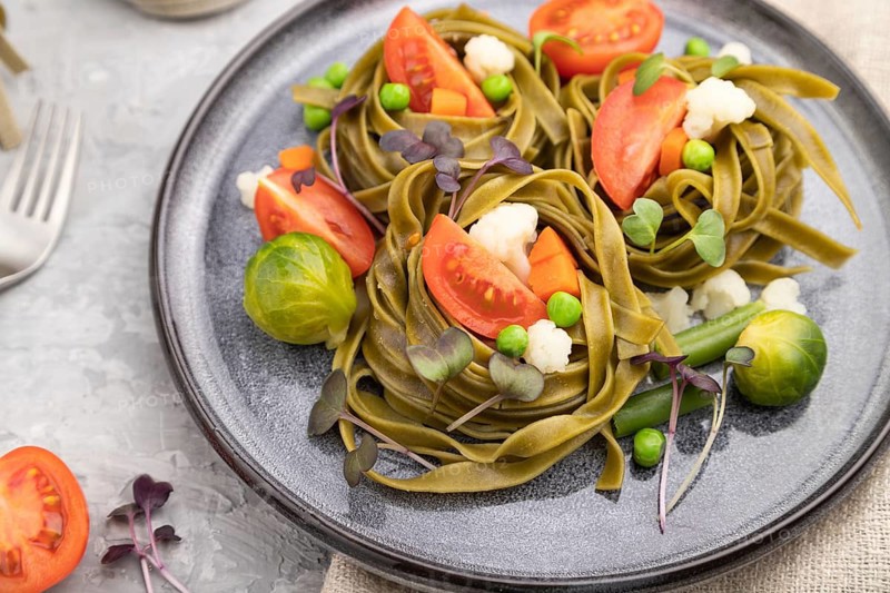 Tagliatelle green spinach pasta with tomato, pea and microgreen sprouts on a gray concrete background and linen textile. Side view, close up, selective focus