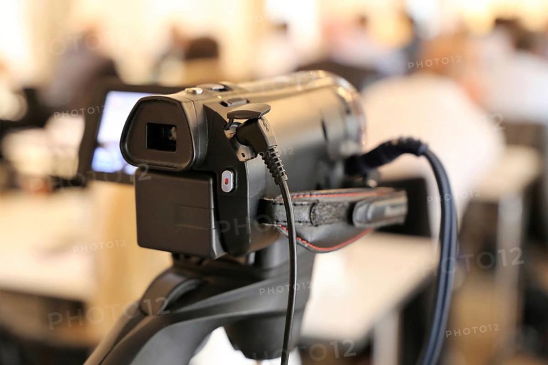 Close-up of a camcorder at a conference or press conference. The guests can be recognised out of focus in the background