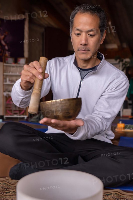 A vertical portrait of a multiracial Latino man with Asian features, holding a Tibetan singing bowl. His expression reflects serenity and spiritual connection
