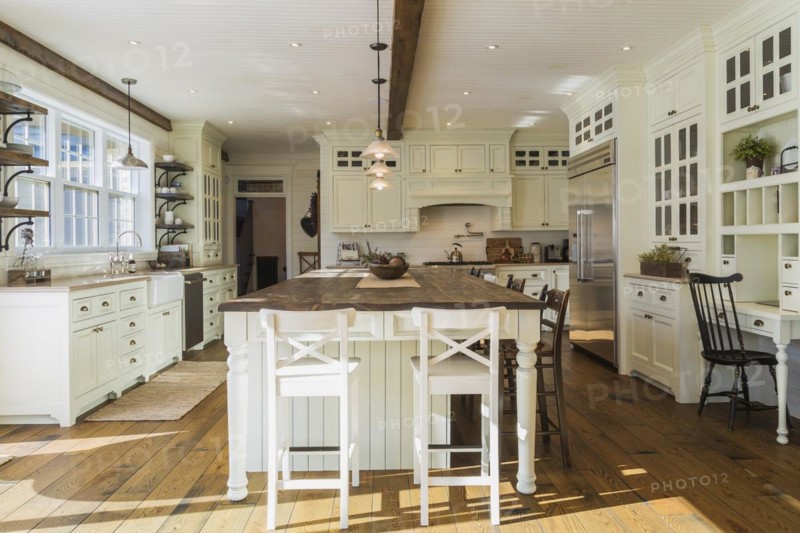 White painted maple wood antique style cabinets with tan coloured quartz countertops, island with brown stained wood plank countertop and high back bar stools in kitchen with stained oak wood floorboards inside country style home, Quebec, Canada, North America
