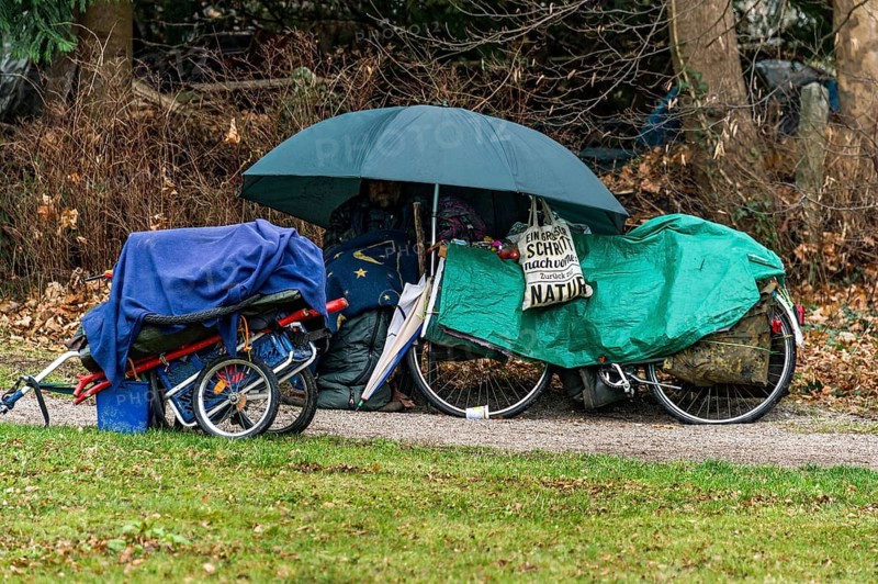 Homeless man in camp on park bench under umbrella with blanket and sleeping bag, bicycle covered with tarpaulin, bicycle trailer with personal belongings, park, autumn, cold, wet, Freising, Upper Bavaria, Bavaria, Germany, Europe
