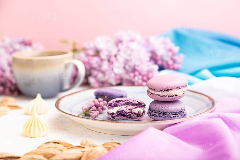 Purple macarons or macaroons cakes with cup of coffee on a white concrete background and magenta-blue textile. Side view, close up, selective focus